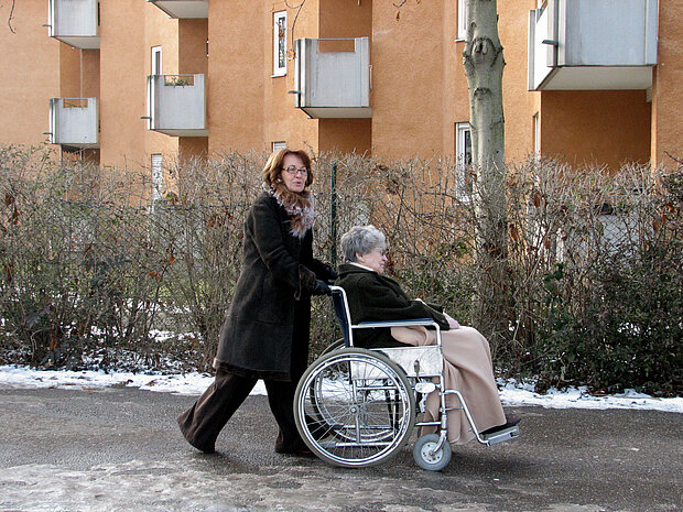 Frau schiebt eine ältere Dame im Rollstuhl einen Weg entlang