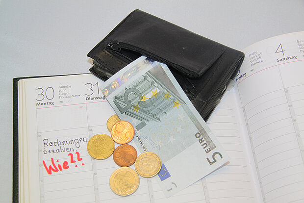 Kalender mit dem Eintrag "Rechnungen bezahlen" und der Notiz "Wie?", darauf eine Geldbörse mit wenigen Euros