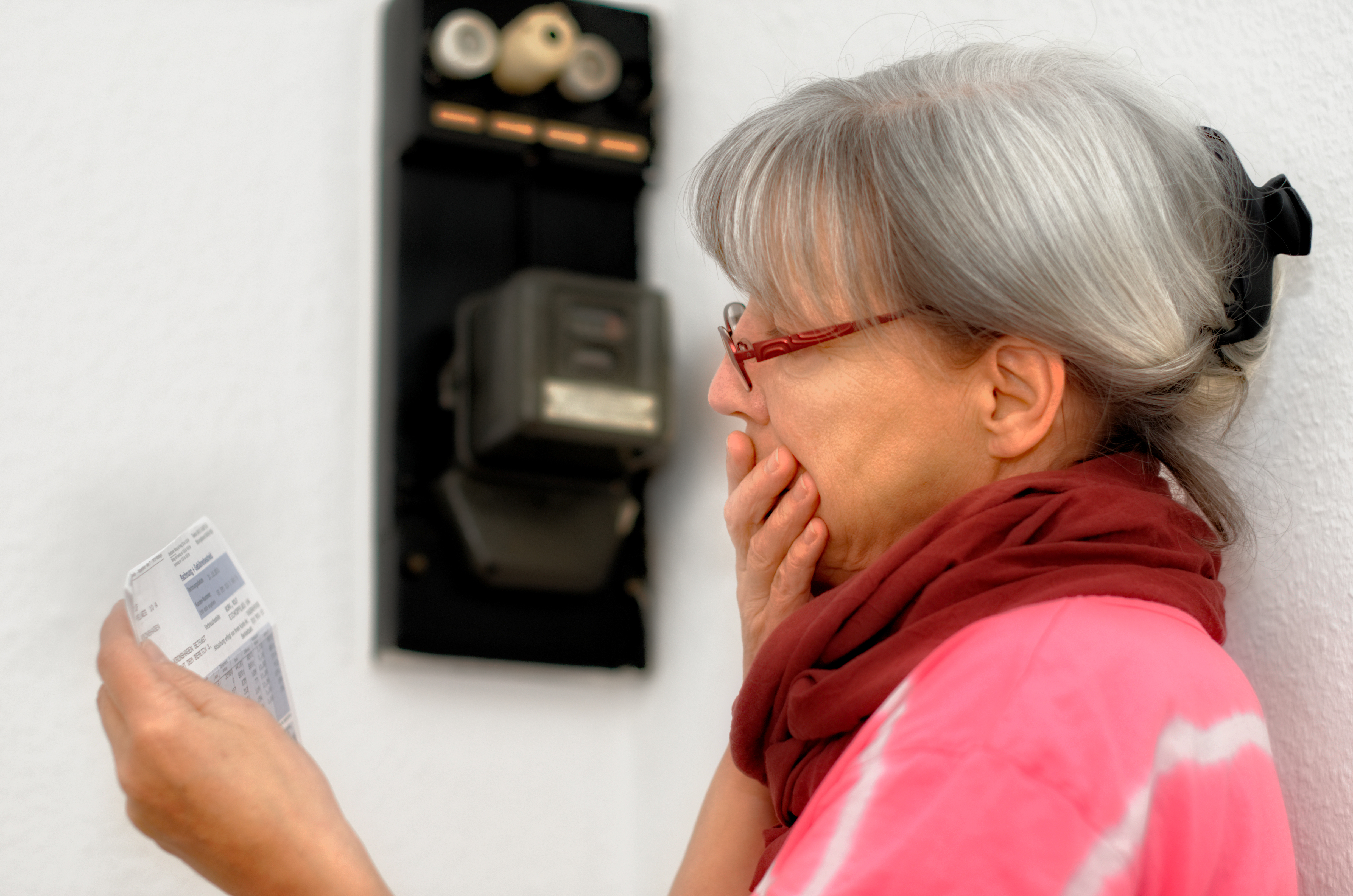 Rentnerin vor Stromzähler erschrickt über Rechnung in ihrer Hand